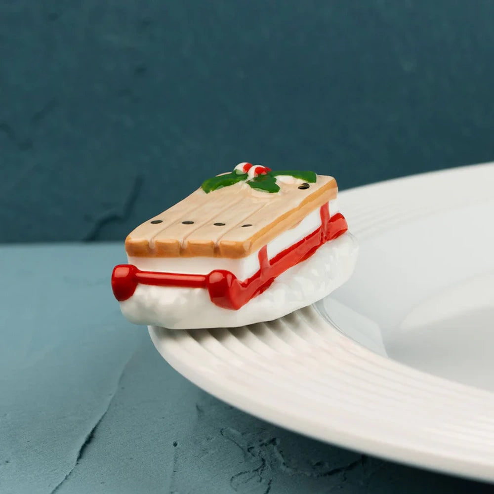 Small ceramic sled figurine with red runners and holly on top, resting on the rim of a white plate.