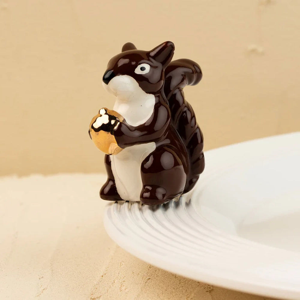 A ceramic squirrel figurine with a glossy brown and white body holding a shiny gold acorn on a white plate.