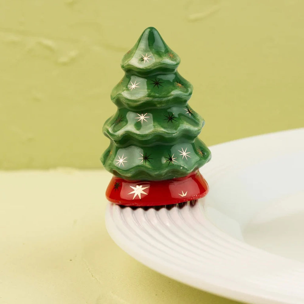 Small ceramic Christmas tree figurine with gold stars and a red base, sitting on the edge of a white plate.
