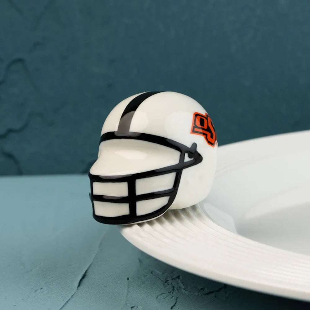 A mini white ceramic football helmet with a black facemask and orange OSU logo, resting on a plate rim.