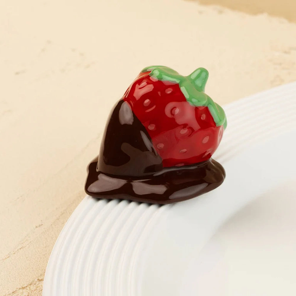 Ceramic chocolate-dipped strawberry figurine resting on the rim of a white textured plate.