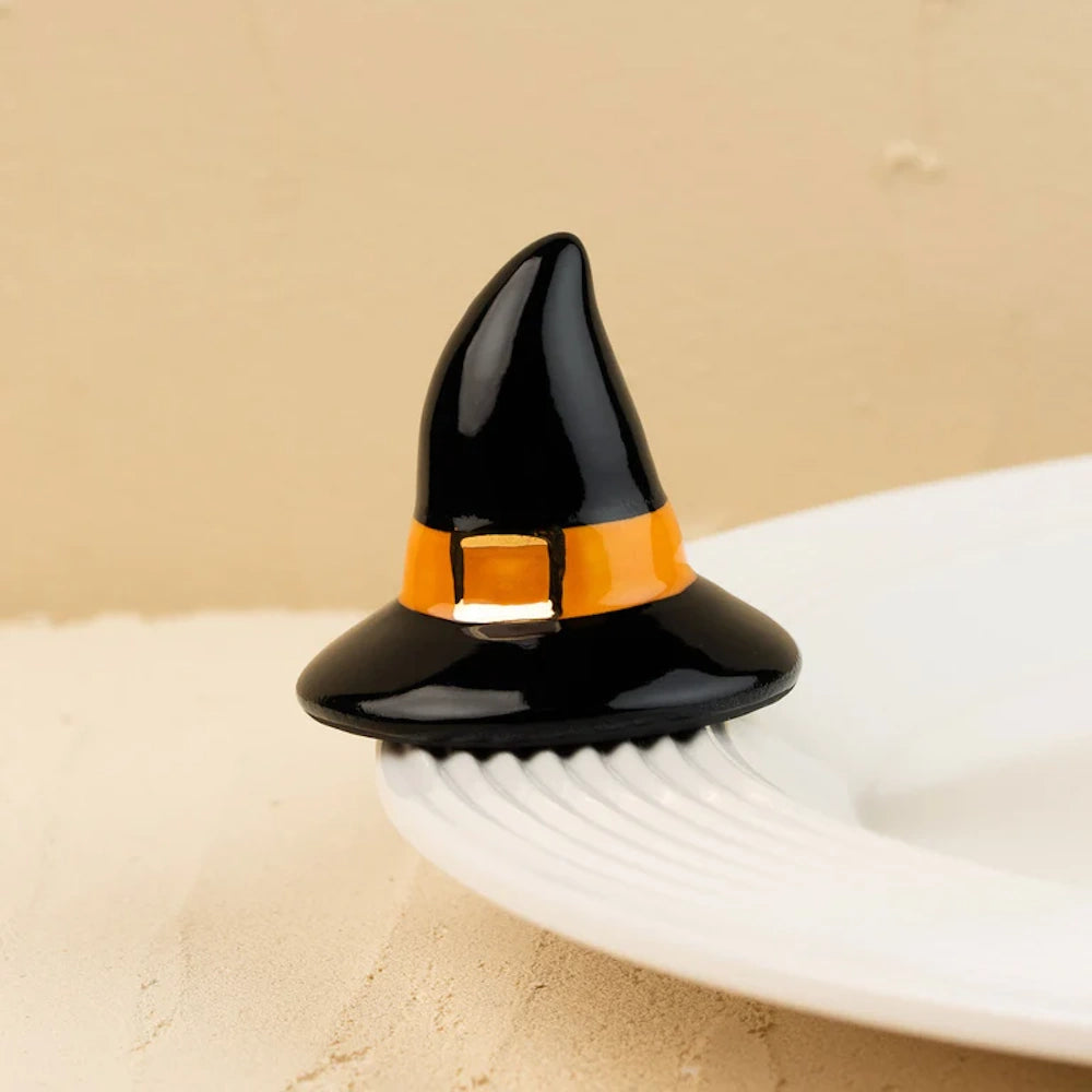 A glossy black ceramic witch hat figurine with an orange band, perched on the rim of a white plate.