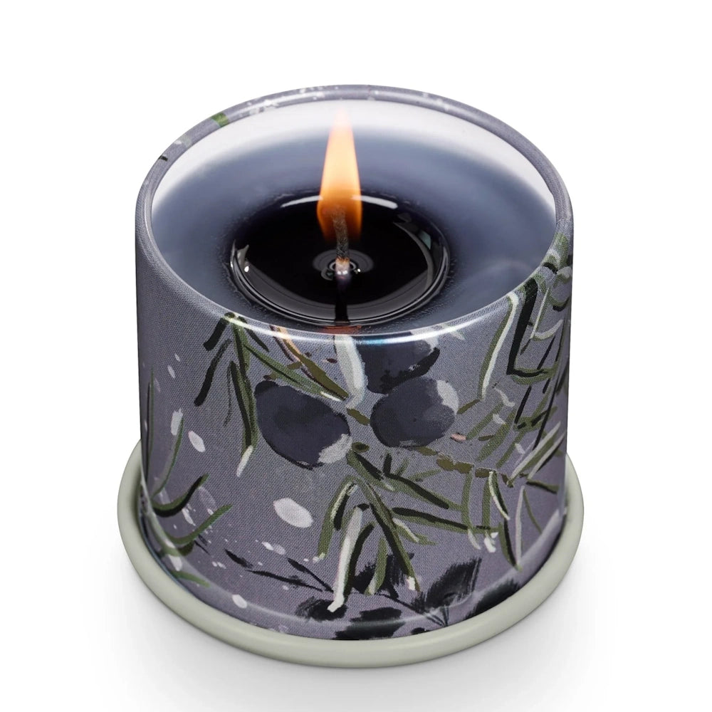 Lit small tin candle with a warm flame, pool of melted wax, and a dark grey botanical evergreen pattern.