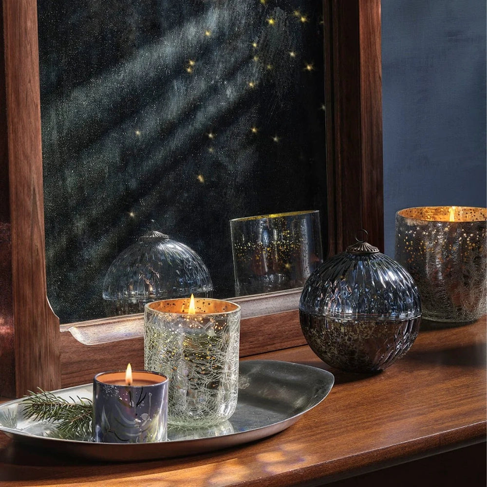 Lit tin candle on a silver tray with mercury glass holders, an ornament, and a mirror reflecting warm light.