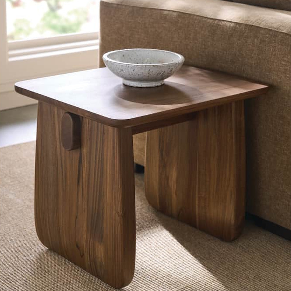 Modern walnut end table styled beside a sofa, featuring a rounded top and sculptural panel leg design.