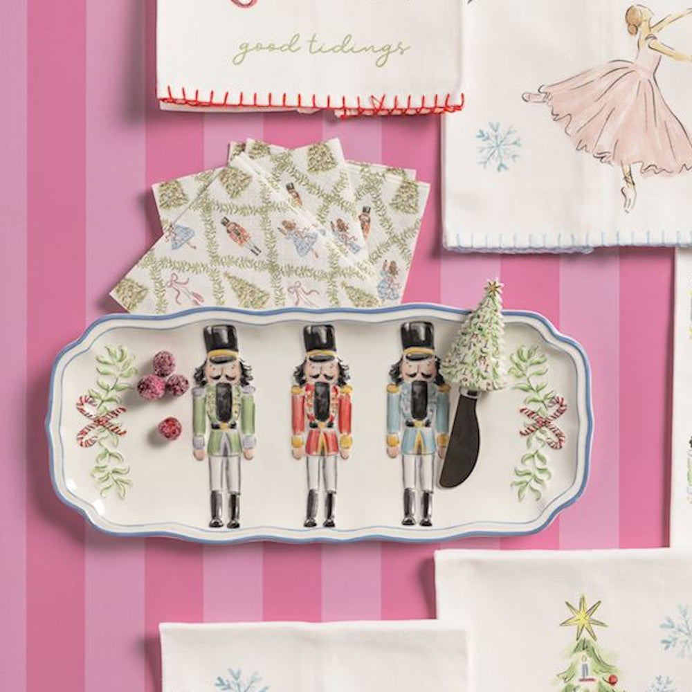 Nutcracker serving platter with tree spreader and patterned napkins set against a pink striped backdrop.