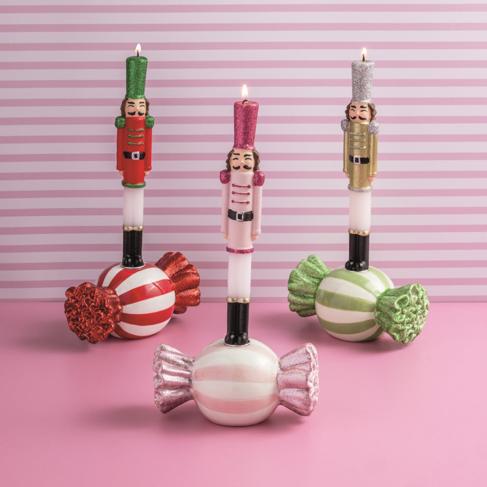 Front view of three lit nutcracker taper candles styled in ceramic candy holders against a pink striped background.