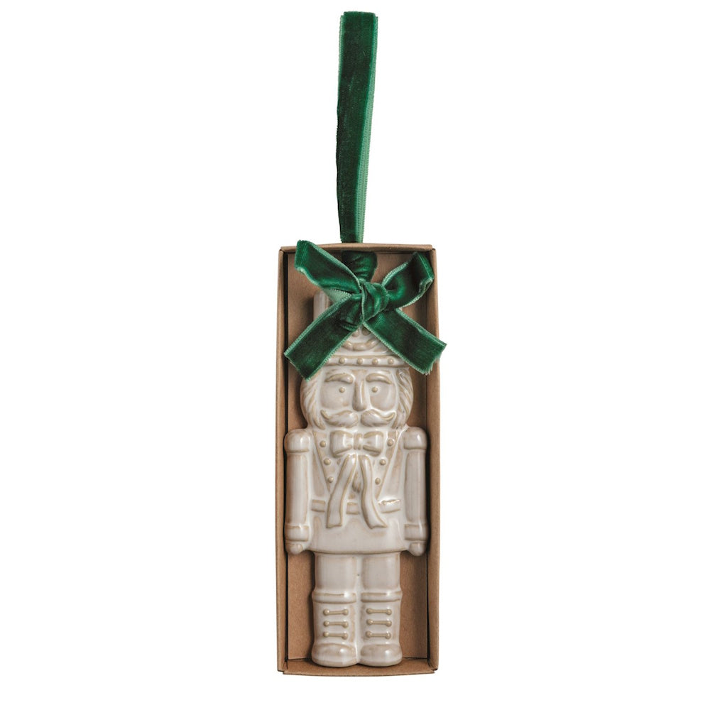 White ceramic nutcracker Christmas ornament in a box with a green velvet bow.
