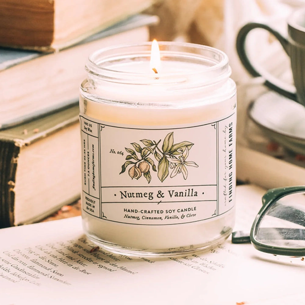 Lit Nutmeg & Vanilla soy candle in a clear glass jar, set against stacked vintage books and eyeglasses.