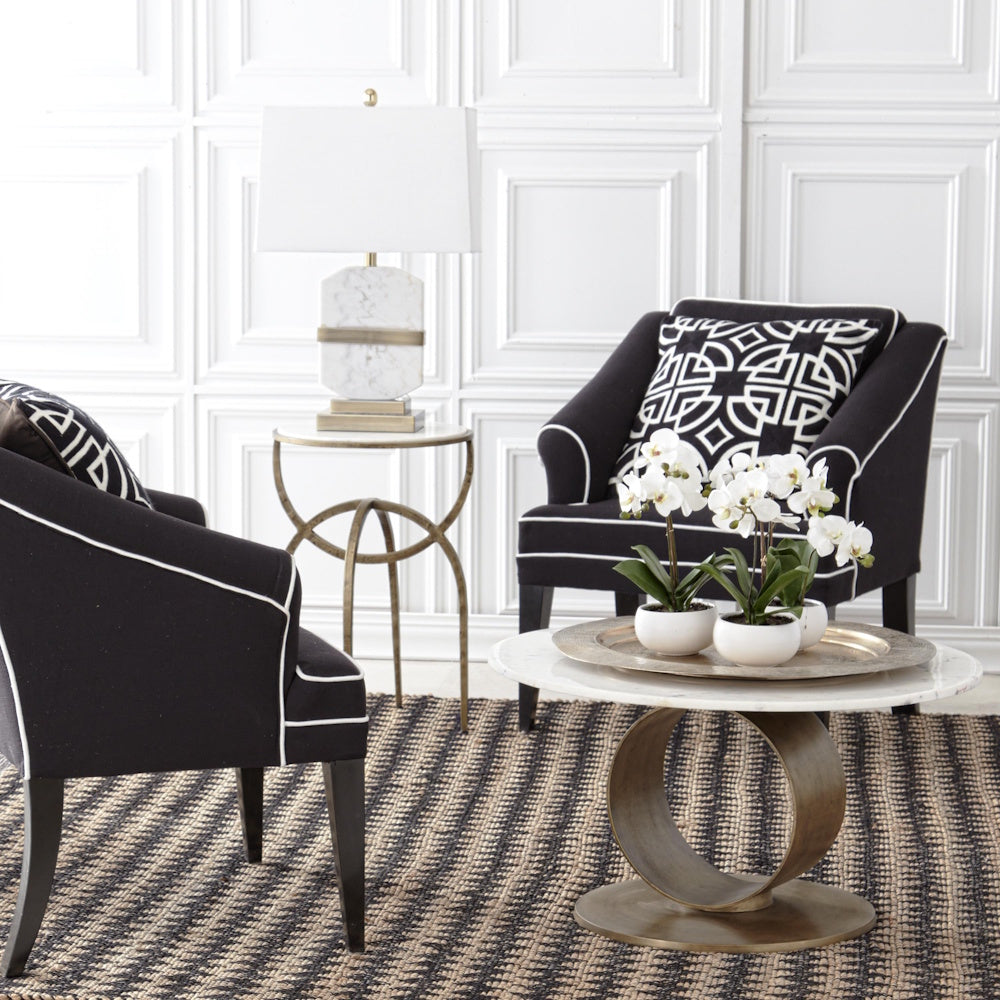 Modern white marble lamp on a gold side table between two black armchairs in a bright, contemporary living room.