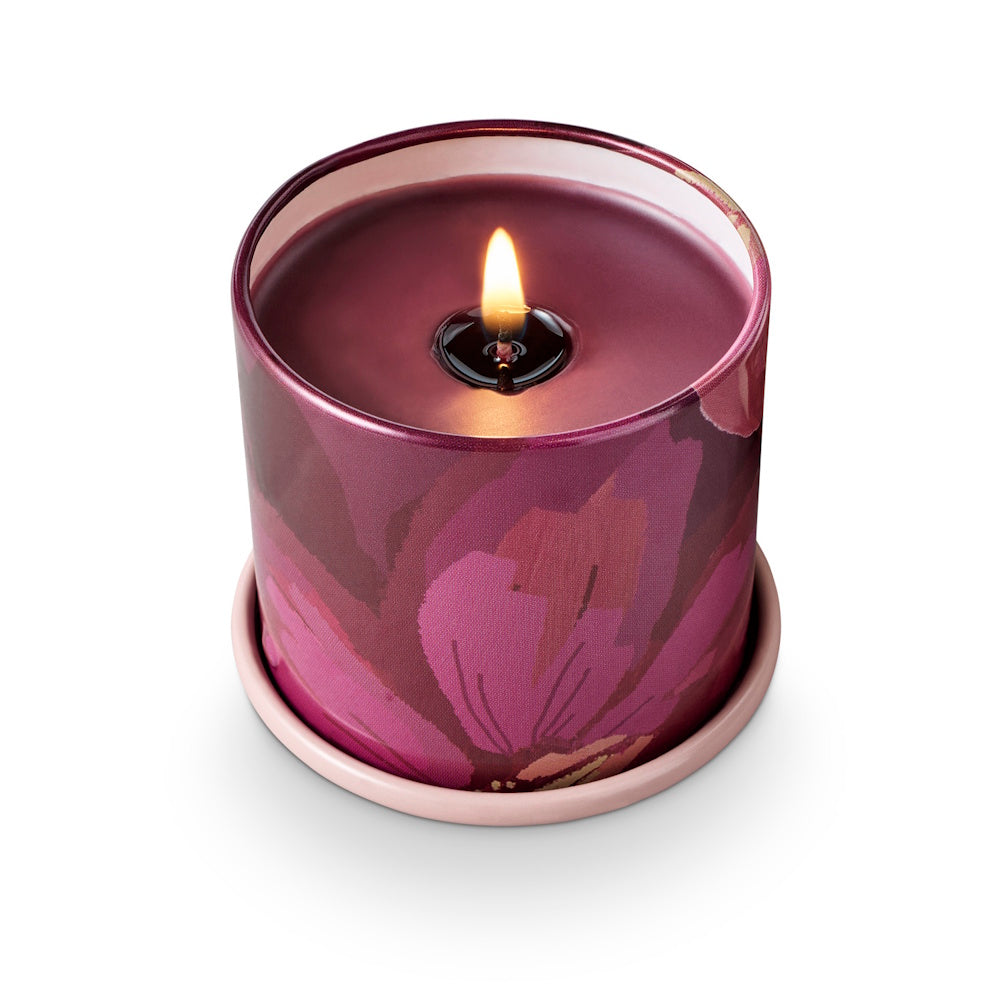 A lit candle with purple wax in a floral pink and plum tin container, resting on a small pink saucer.