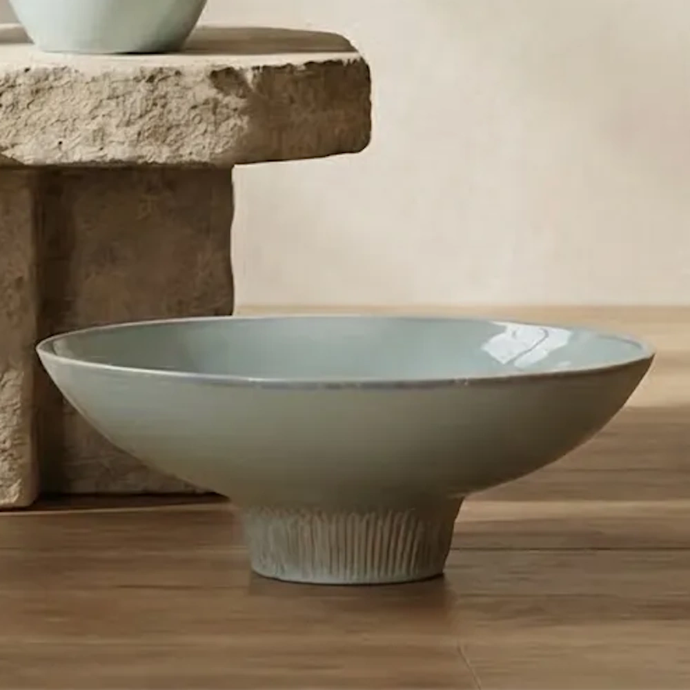 Light blue ceramic pedestal bowl resting on a wooden surface next to a stone pedestal.