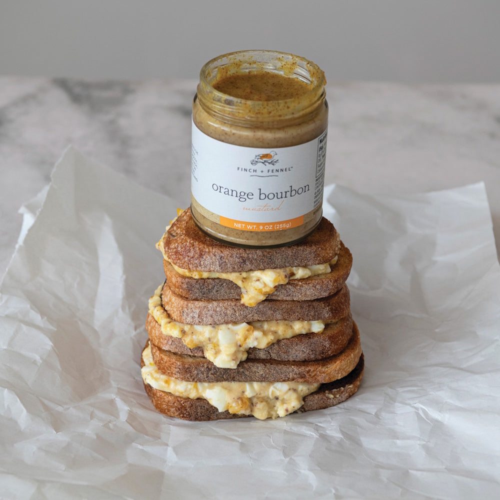 An open jar of Finch + Fennel Orange Bourbon Mustard sits on a stack of egg salad sandwiches on white paper.