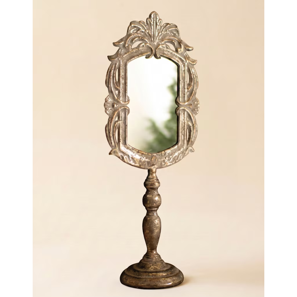 Ornate rustic metal vanity mirror on a tall pedestal base against a solid beige background.