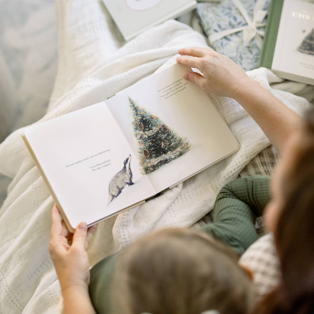A person reading a Christmas storybook with a badger and decorated tree illustration to a child.