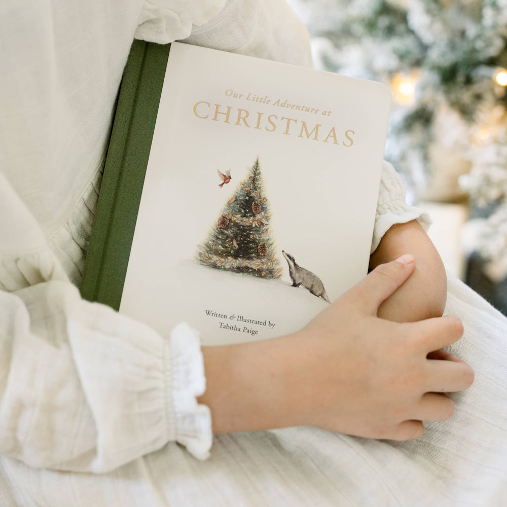 A child holding "Our Little Adventure at Christmas" book by Tabitha Paige, featuring a watercolor tree and badger.