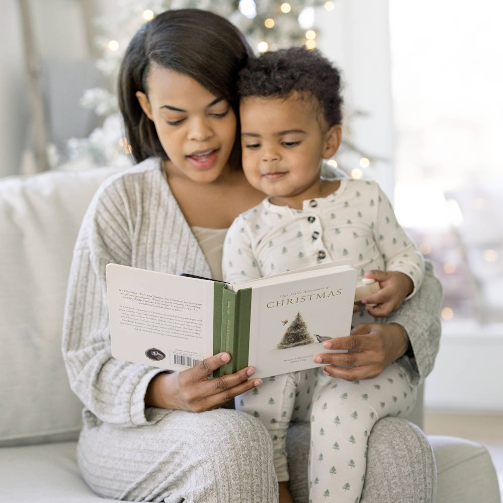 Woman and child in pajamas reading "Our Little Adventure at Christmas" book together on a sofa with holiday lights.