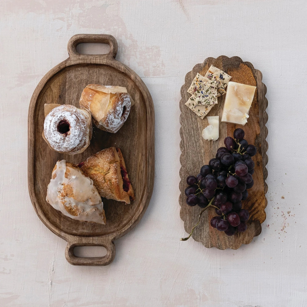 An oval wooden tray with pastries alongside a scalloped tray with cheese, crackers, and grapes on a light surface.