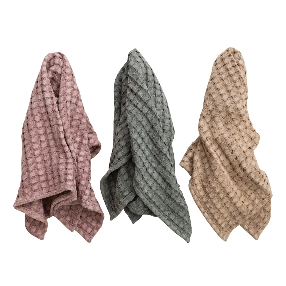 Three stonewashed cotton waffle-weave tea towels in dusty rose, sage green, and tan hanging against a white background.