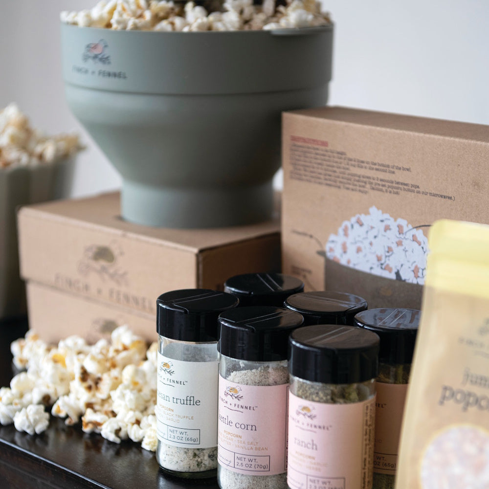 An assortment of Finch + Fennel popcorn seasonings in front of a green silicone popcorn popper and boxes.