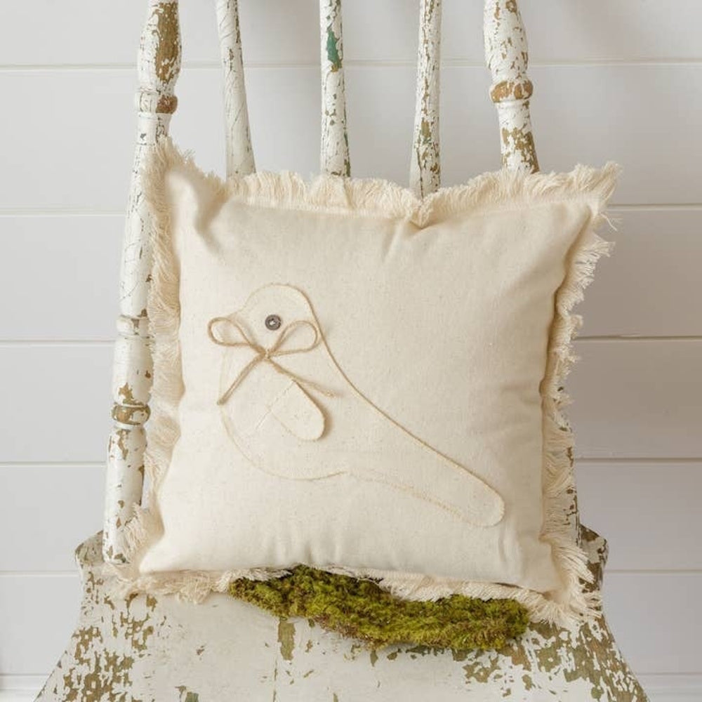 A cream-colored fringed pillow with a fabric bird patch and twine bow, sitting on a rustic chair.