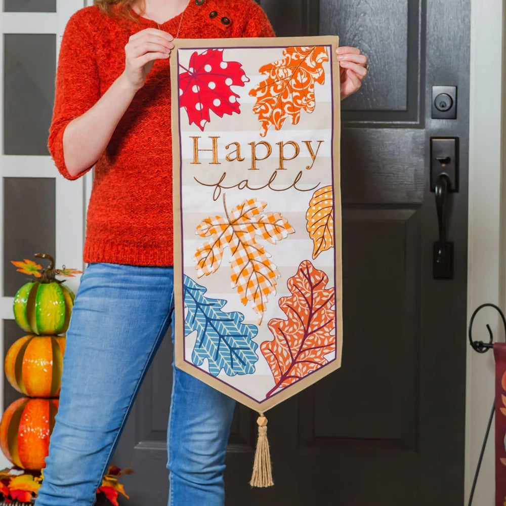 Person holding a "Happy fall" wall banner with colorful patterned autumn leaves near a front door.