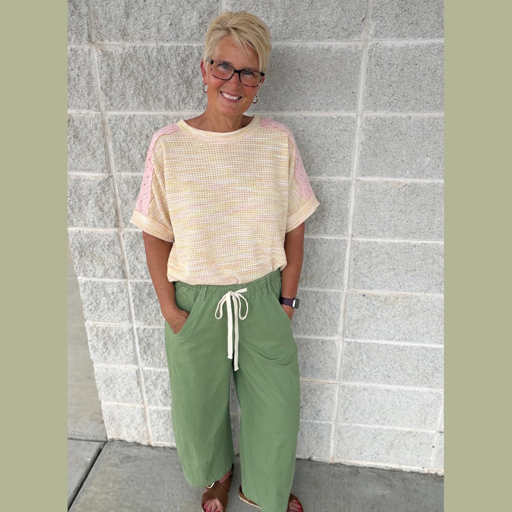 Woman in a peach and lemon eyelet top with green wide-leg pants and brown sandals against a stone wall.