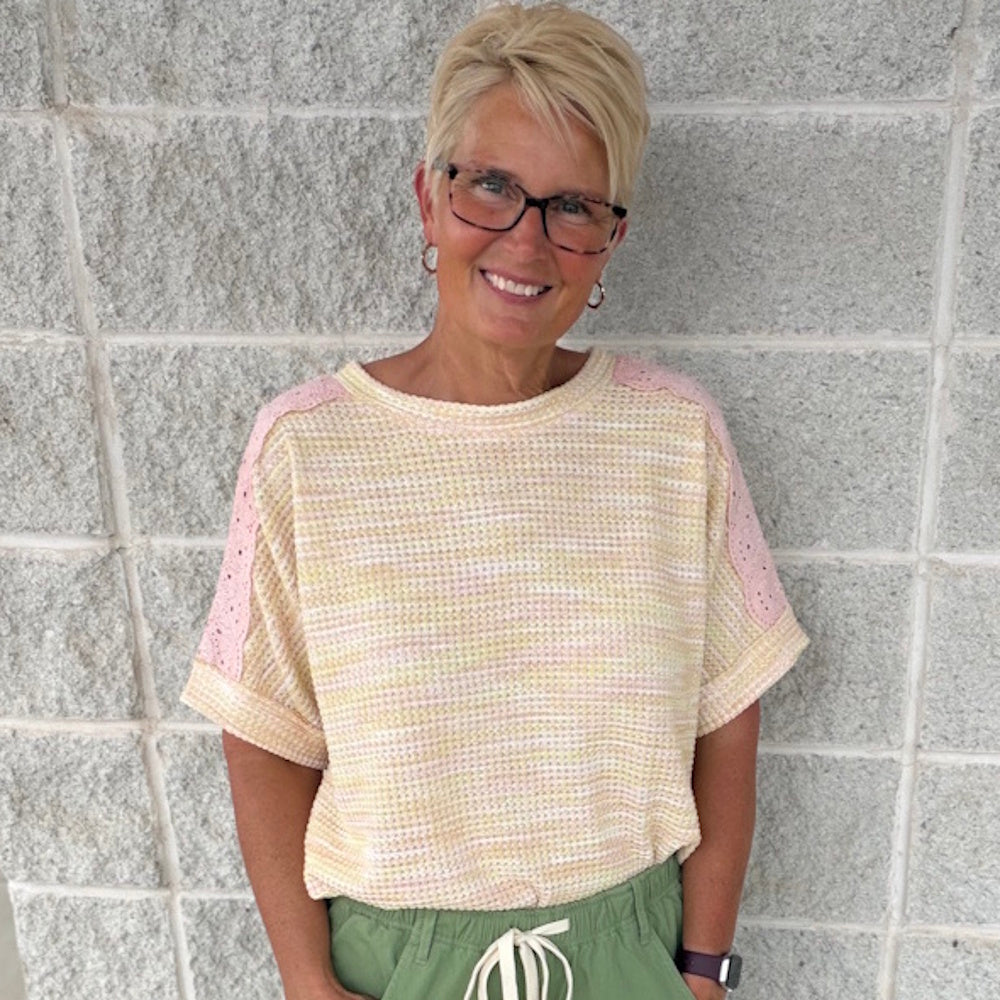 Smiling woman with short blonde hair and glasses wearing a yellow knit top with pink lace shoulders.
