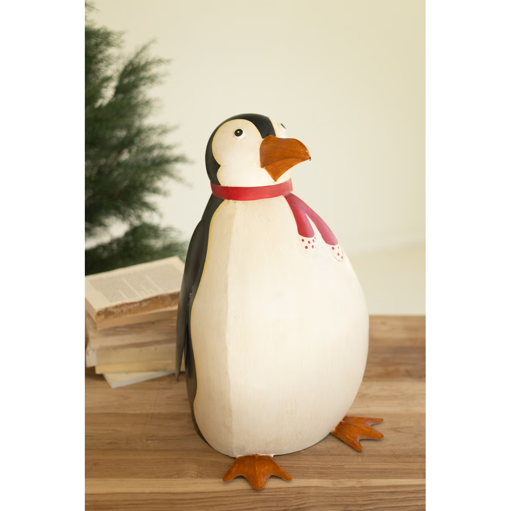 Large painted metal penguin sculpture wearing a red scarf placed on a rustic wooden table.