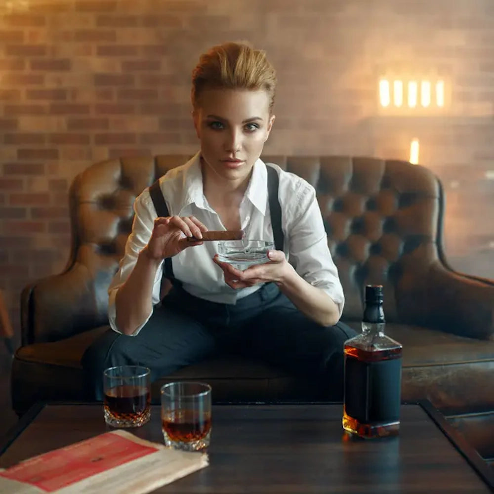 Woman in a white shirt and suspenders sitting on a leather sofa holding a cigar over an ashtray near whiskey.