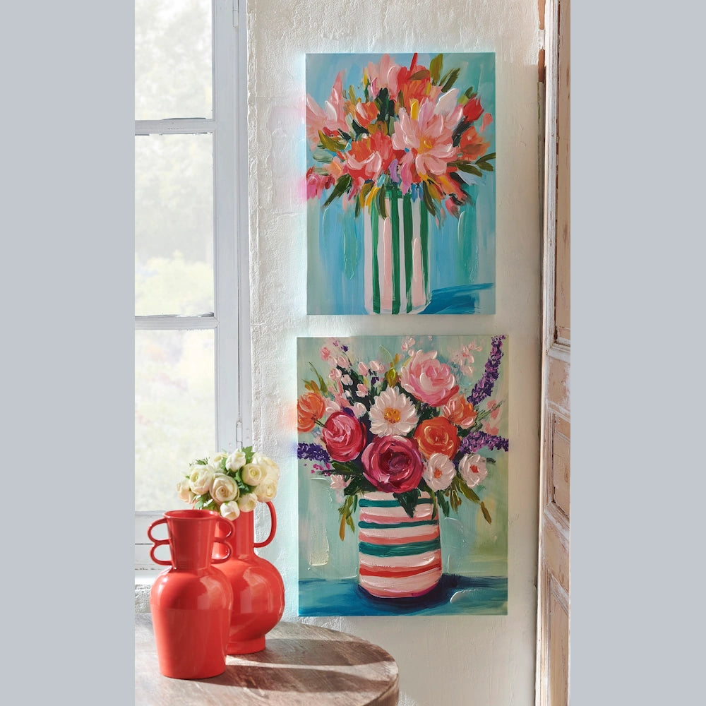 Two colorful floral oil paintings on a white wall next to a window and two bright red vases on a wooden table.