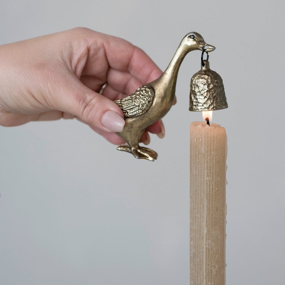 A hand holds a brass duck figurine carrying a small bell snuffer over a lit textured taper candle.