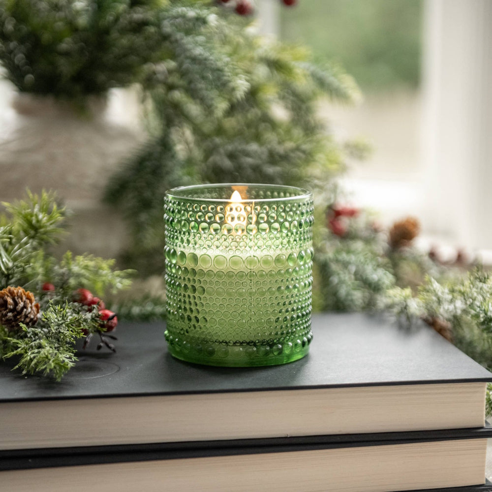 Lit candle in a green dotted glass holder sitting on stacked books with pine greenery nearby.