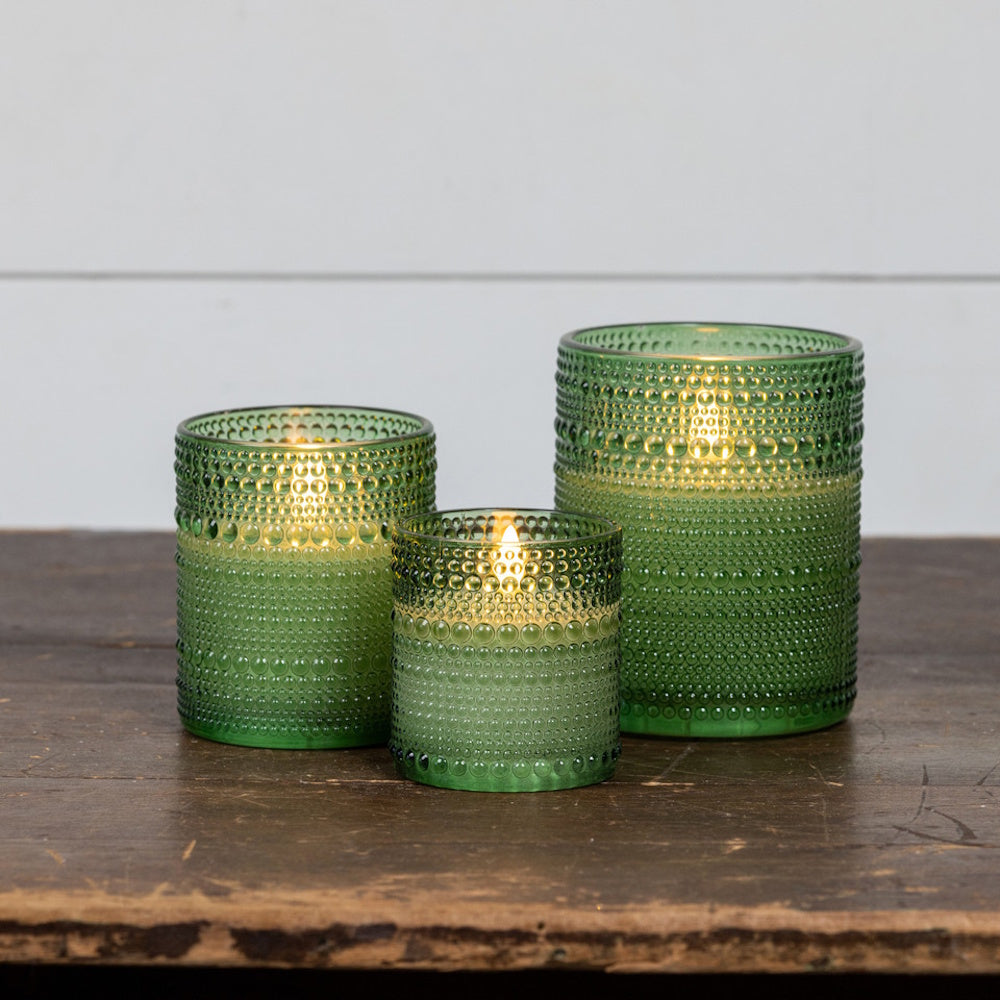 Three glowing green textured glass candle holders in varying sizes resting on a rustic wooden table.