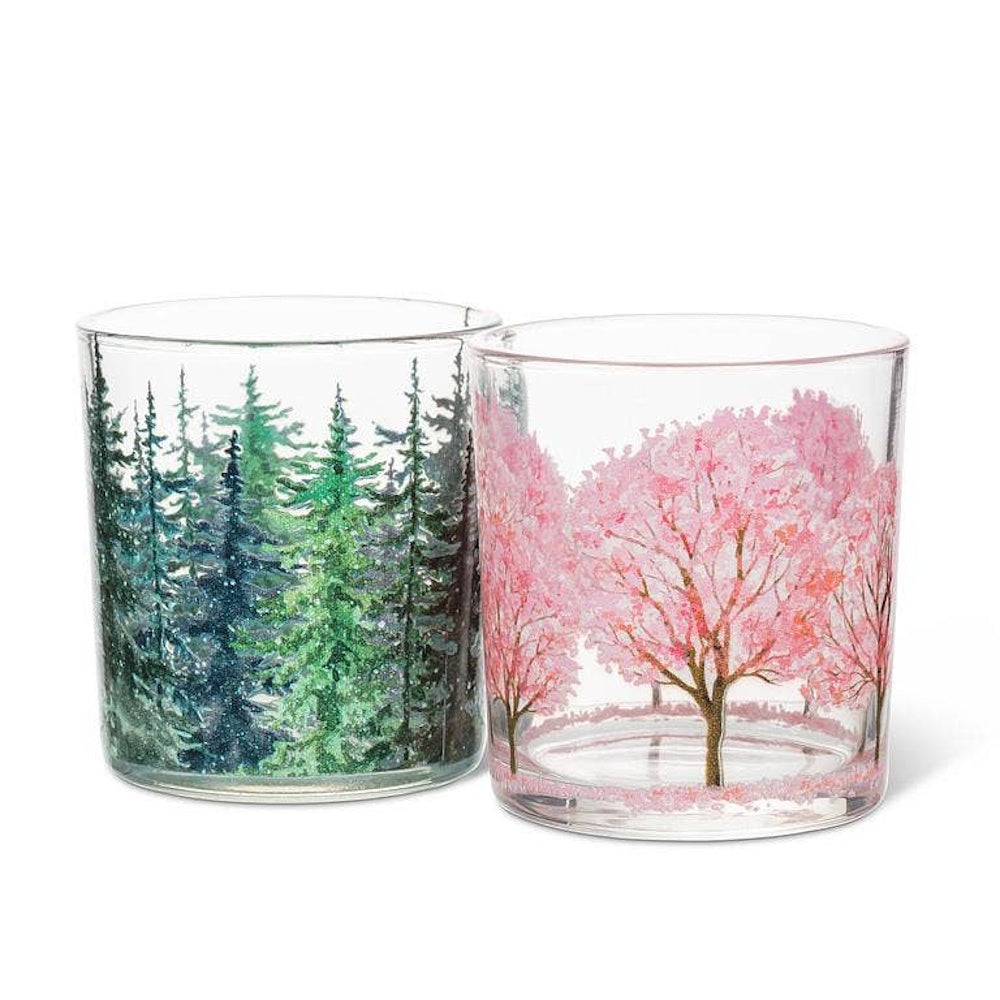 Two glass votive holders, one featuring green pine trees and the other pink cherry blossom trees.