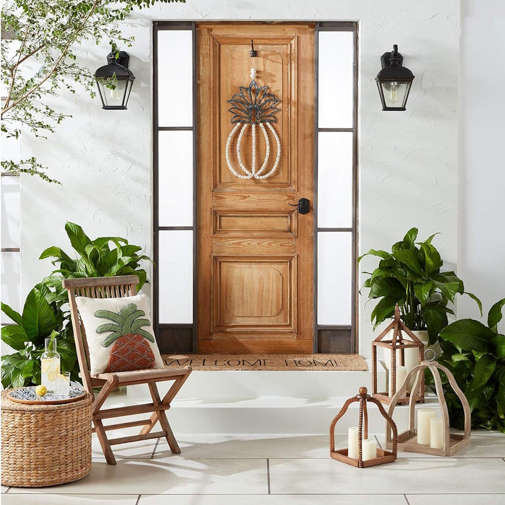 A front porch with a pineapple pillow on a chair, a pineapple wreath on the door, lanterns, and plants.