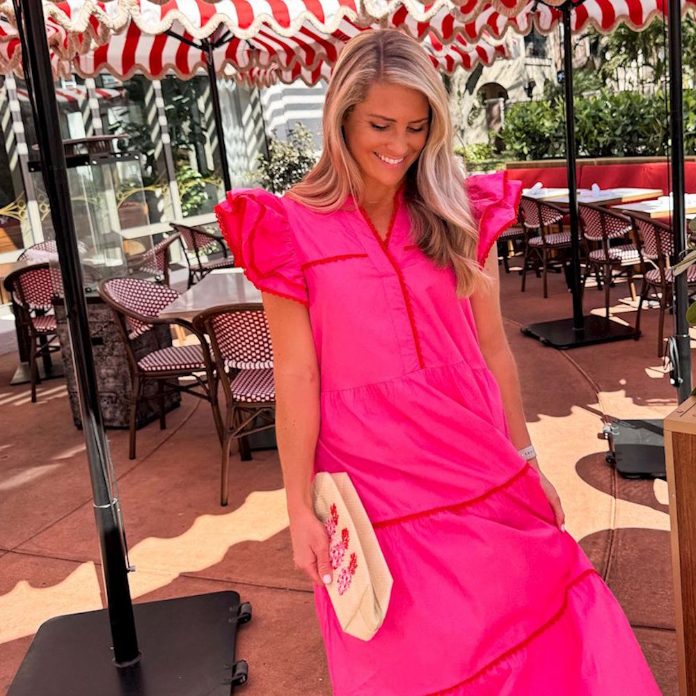 Smiling woman in a bright pink tiered dress holding a woven clutch at an outdoor cafe with striped umbrellas.
