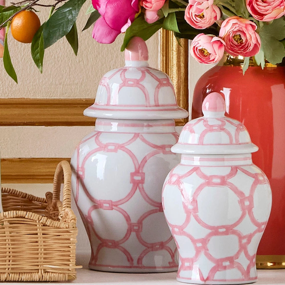 Two white ginger jars with a pink trellis pattern stand next to a wicker basket and pink flowers in a gold vase.