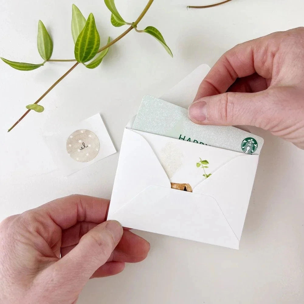 Hands placing a Starbucks gift card into a small white envelope alongside a tiny card.
