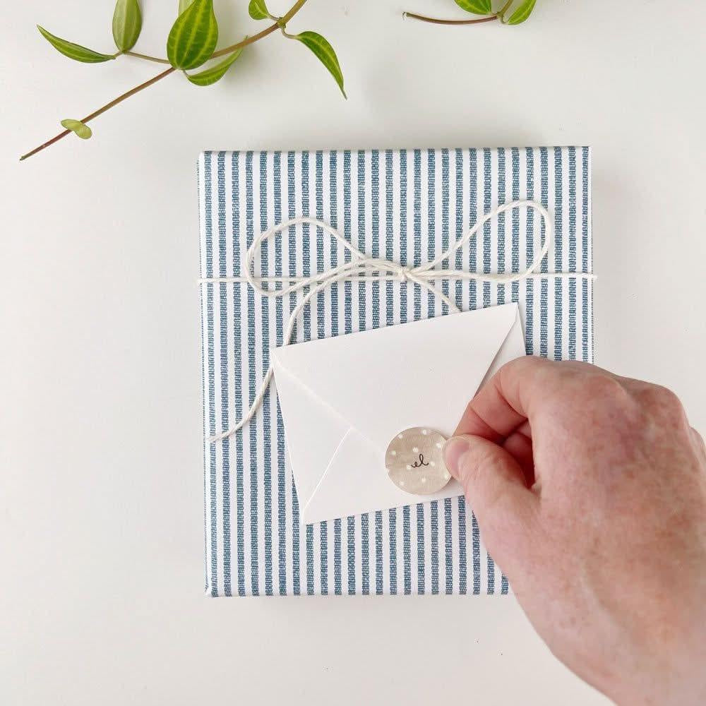 Hand applying a round sticker to seal a mini white envelope on a blue striped gift package.