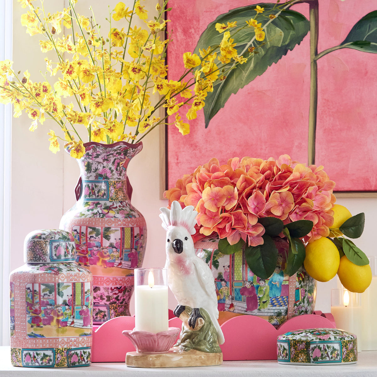 Decorative scene with vibrant yellow and orange flowers in ornate vases. A white porcelain parrot, lit candle, lemons, and pink backdrop complete the setting.