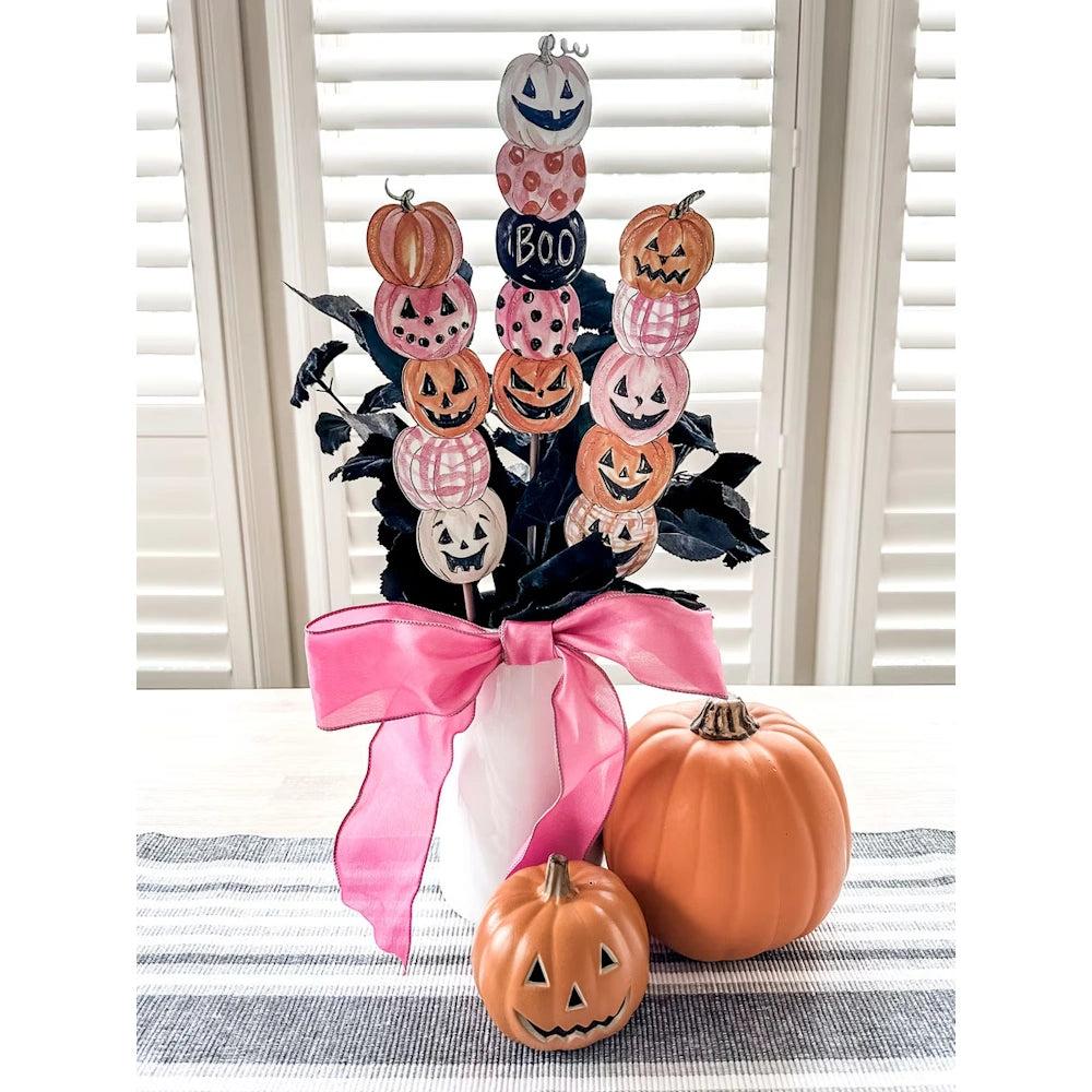 Pumpkin skewers with jack-o'-lanterns in a white vase tied with a pink ribbon, next to orange pumpkins.