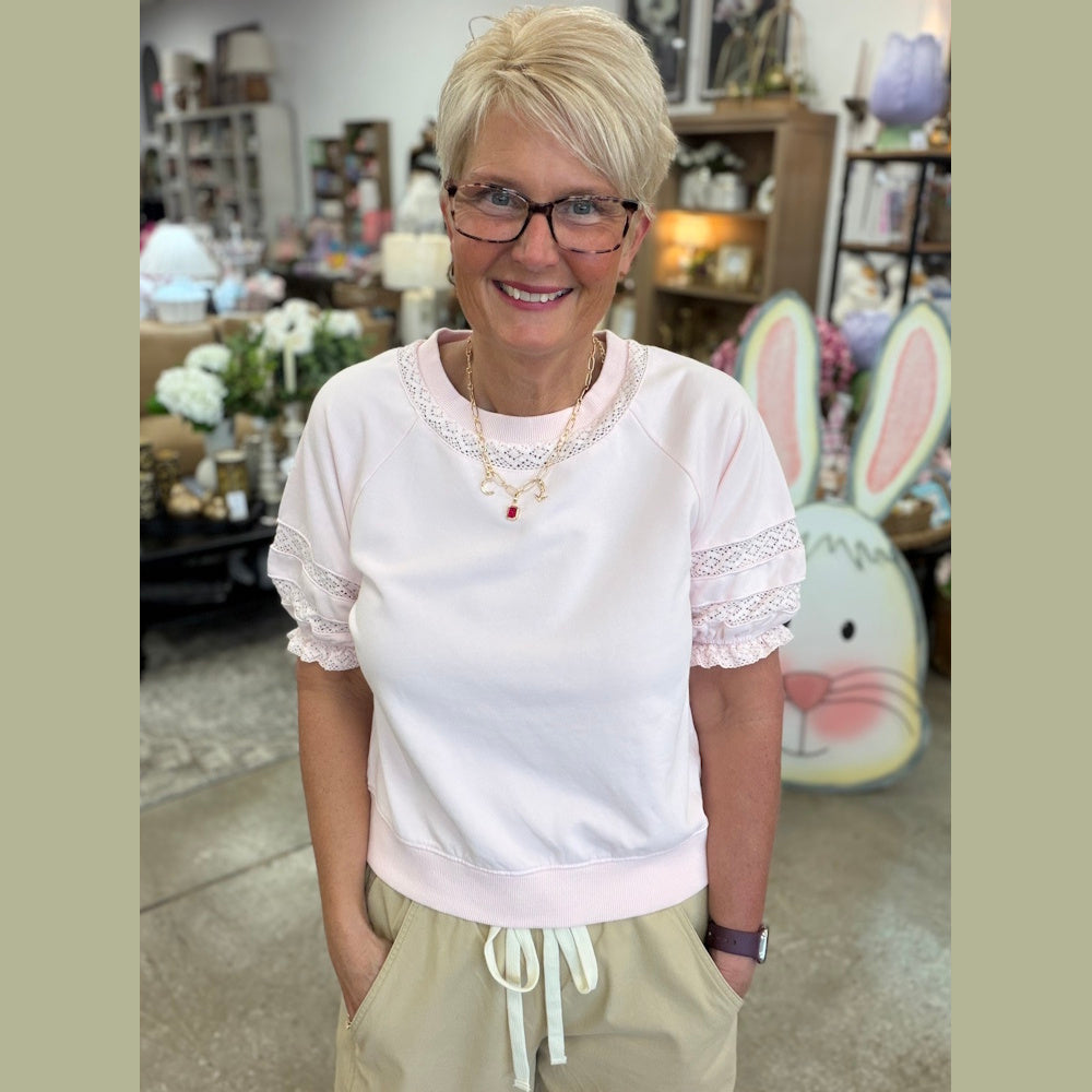 A person with short blonde hair and glasses wearing a pink puff-sleeve top with lace details and khaki pants.