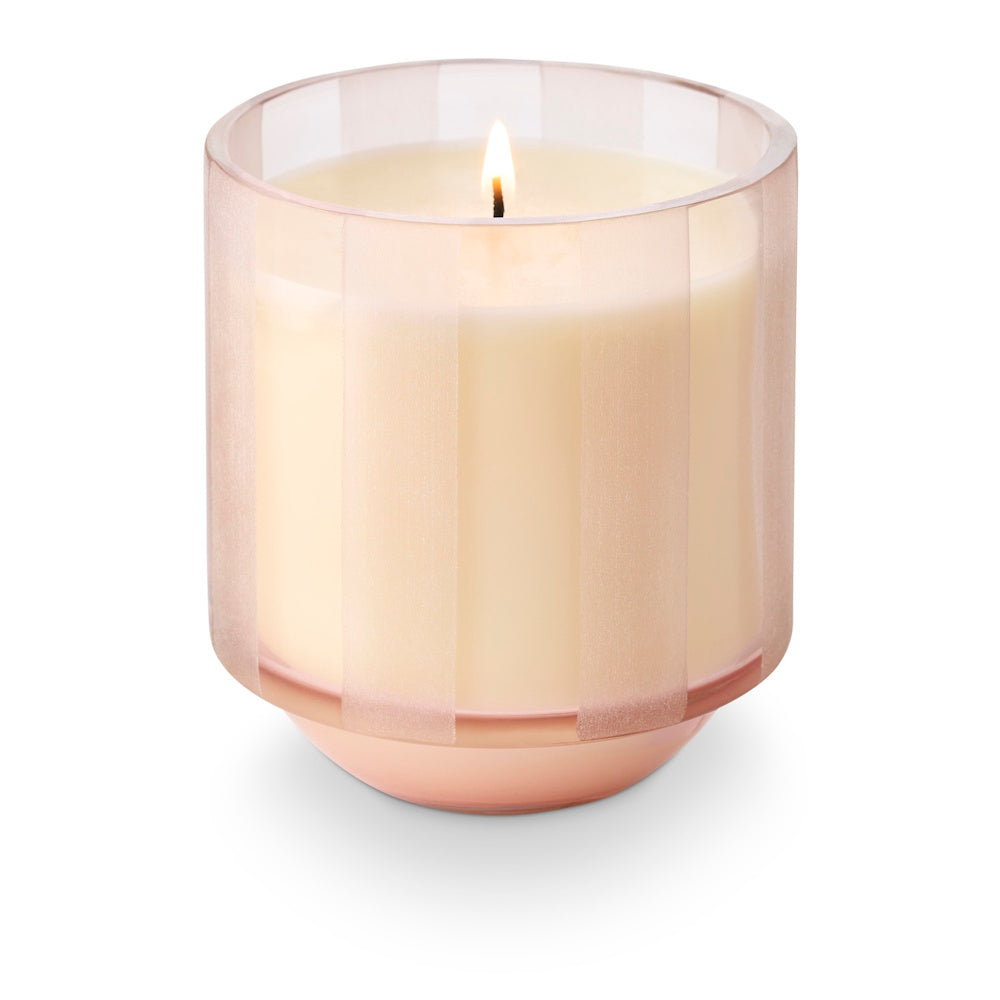 A lit candle in a translucent pink etched-stripe glass jar with a rounded base.