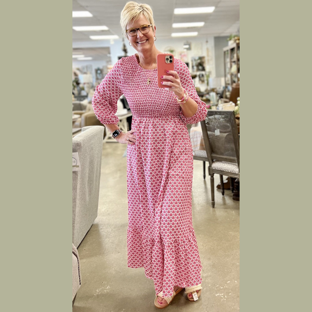A smiling woman in a boutique takes a selfie wearing a pink, patterned, smocked-waist maxi dress.