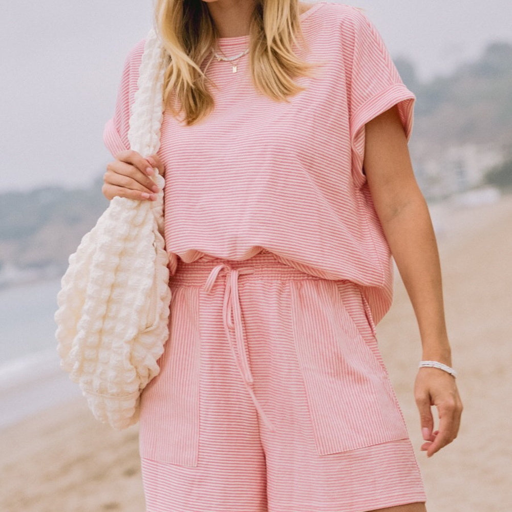 Woman wearing a pink and white striped top and shorts lounge set on a beach while carrying a textured tote bag.