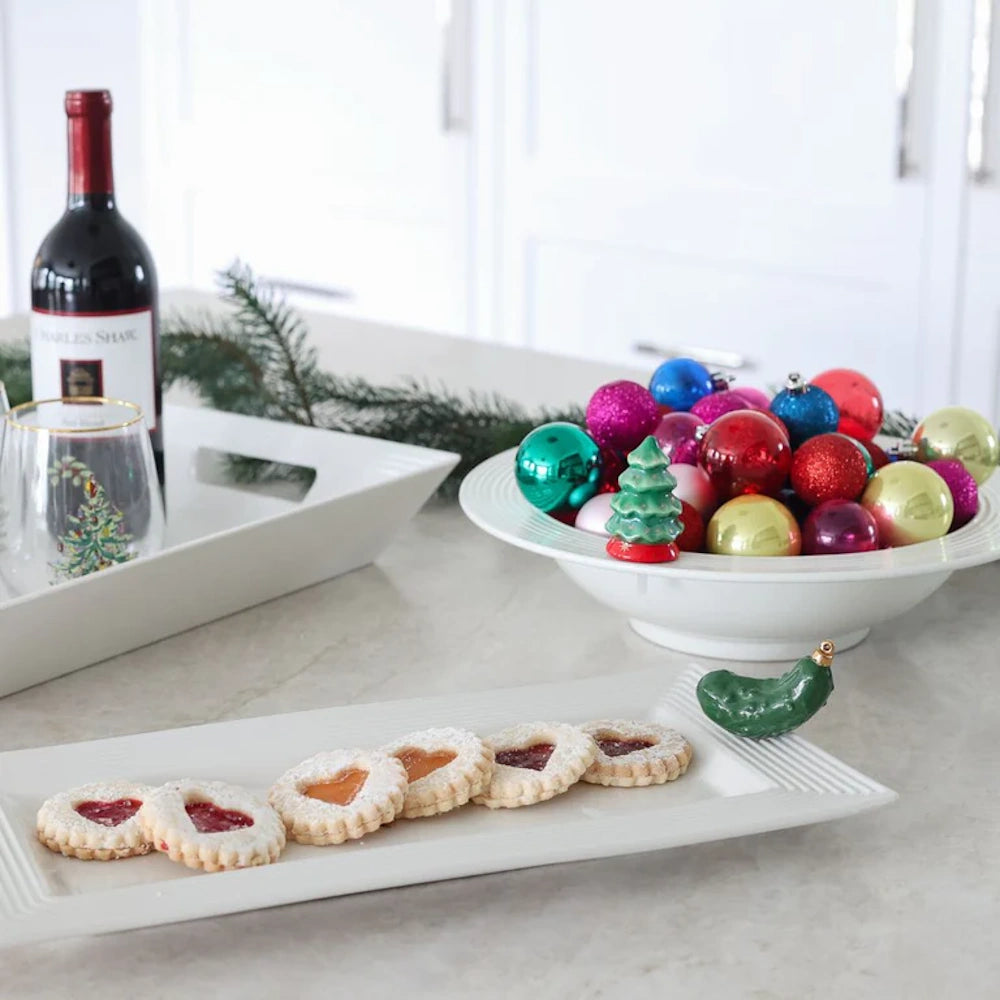 White tray of heart cookies with a ceramic pickle figurine on the rim, next to Christmas decor.