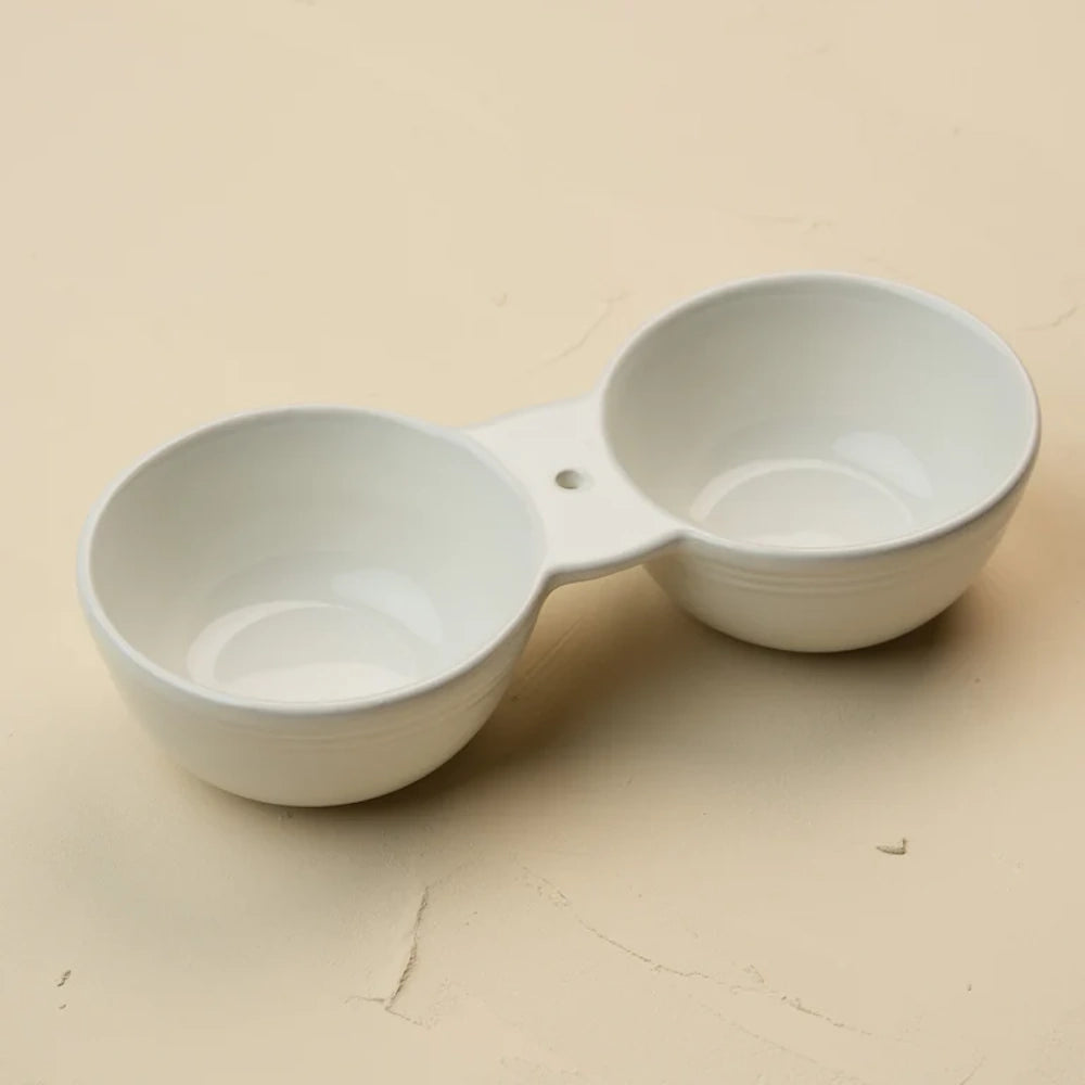A white ceramic double dipping bowl with a central connecting bridge and small hole, on a tan surface.