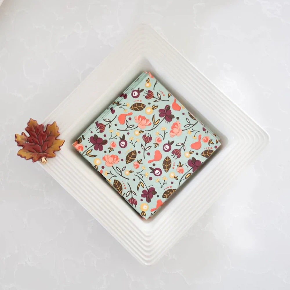 A square white ceramic napkin holder filled with floral napkins and adorned with a ceramic autumn leaf.