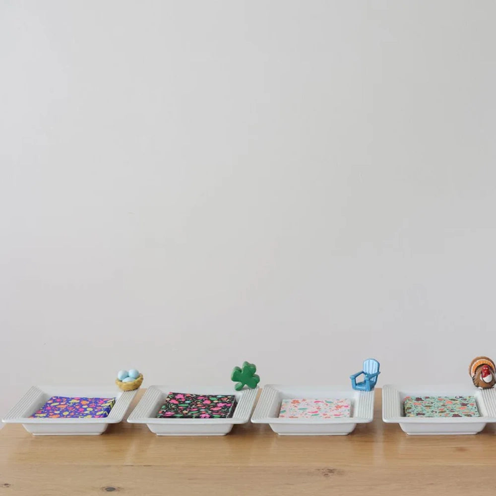 Four white square dishes lined up on a wooden table, each holding napkins and decorated with a small figurine.